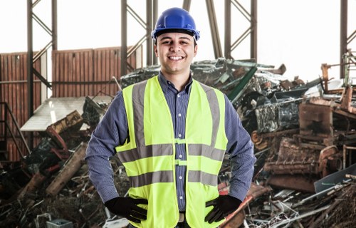 A smiling male worker wearing a blue safety helmet, a high-visibility yellow vest, a long-sleeved blue shirt, and black gloves stands inside a large industrial or warehouse setting. Behind him, there is a pile of various debris and scrap materials, including metal parts, wooden fragments, and other waste objects, which appear to be in the process of disposal or recycling. The background shows structural elements such as metal beams and a partially visible roof, indicating an indoor environment used for waste management or rubbish removal activities. The worker's confident stance and the organized environment reflect professional rubbish collection and waste clearance services, likely undertaken by companies such as Commercial Waste Bracknell in the local area near Bracknell or the surrounding postcodes.