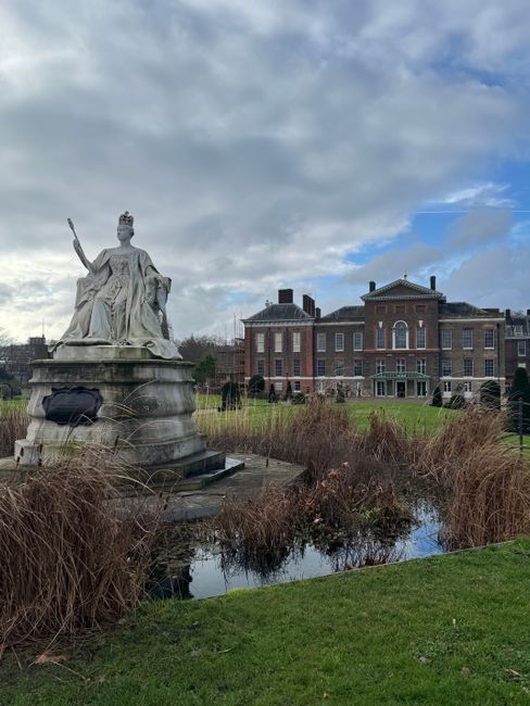 A large stone statue of a regal seated female figure holding a scepter and wearing a crown, situated on a square pedestal with a black bag resting against it, positioned in a landscaped outdoor area f