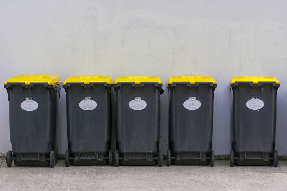 A large green wheeled rubbish bin filled with several red and black plastic bags and a folded cardboard box, situated outdoors against a dark brick wall. The bin appears to contain waste materials typ